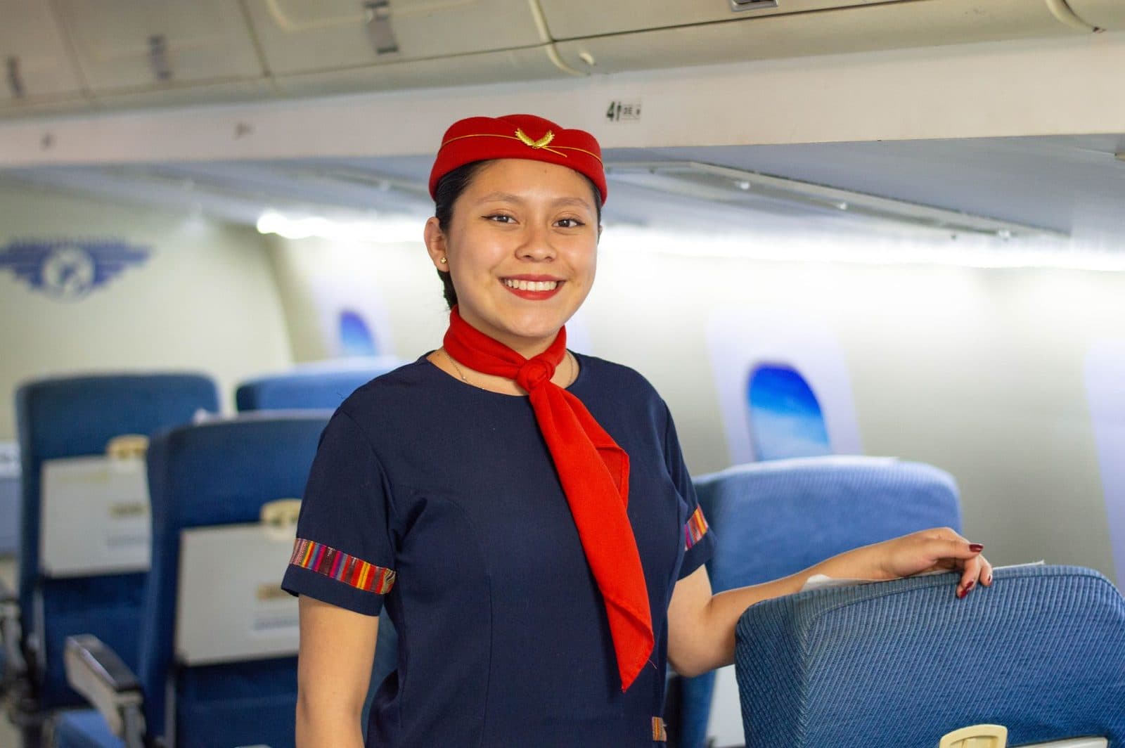 Hôtesse de l'air souriante en uniforme bleu marine, calot et foulard rouges, dans la cabine d'un avion