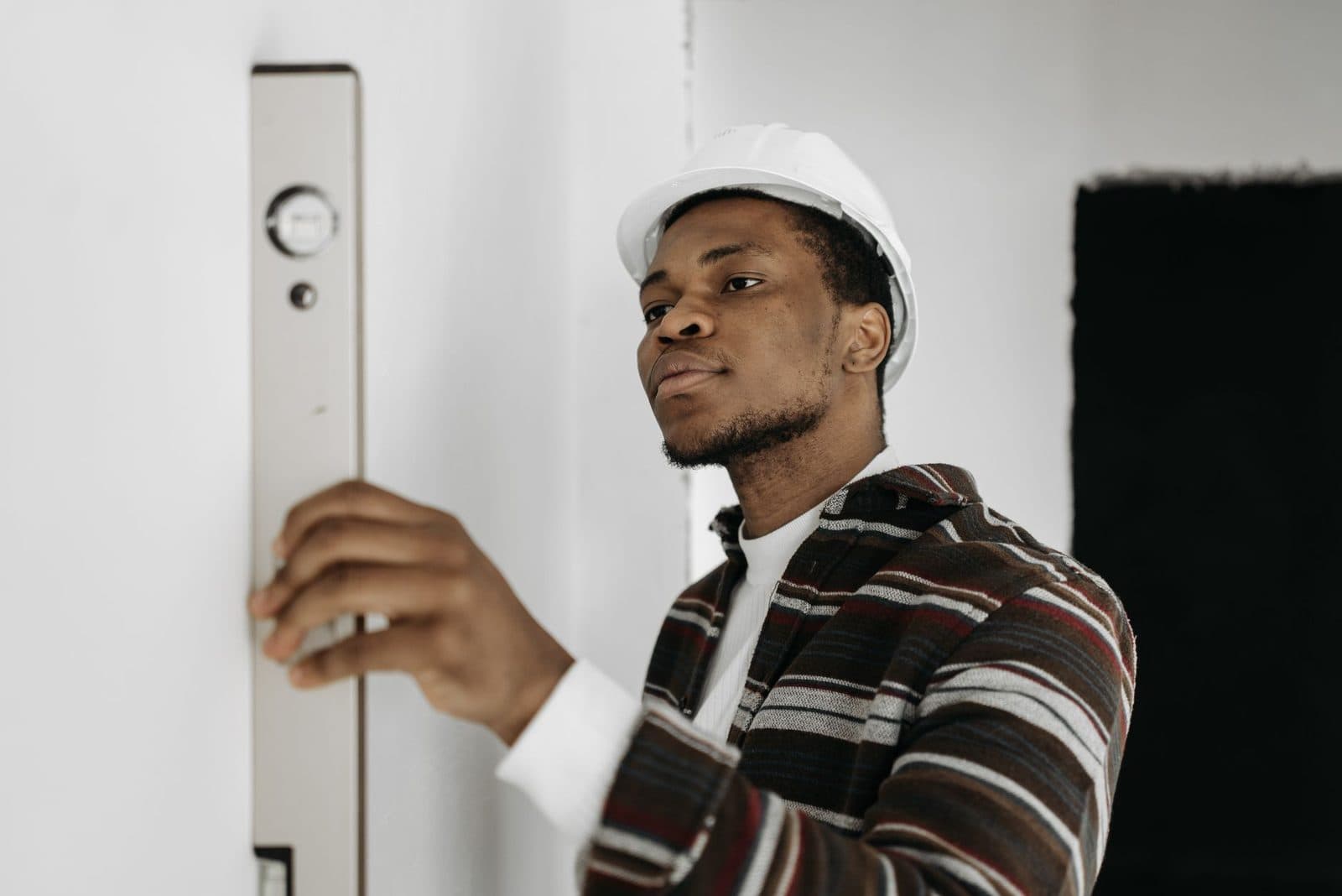 Jeune homme avec casque de chantier blanc utilisant un niveau à bulle contre un mur blanc