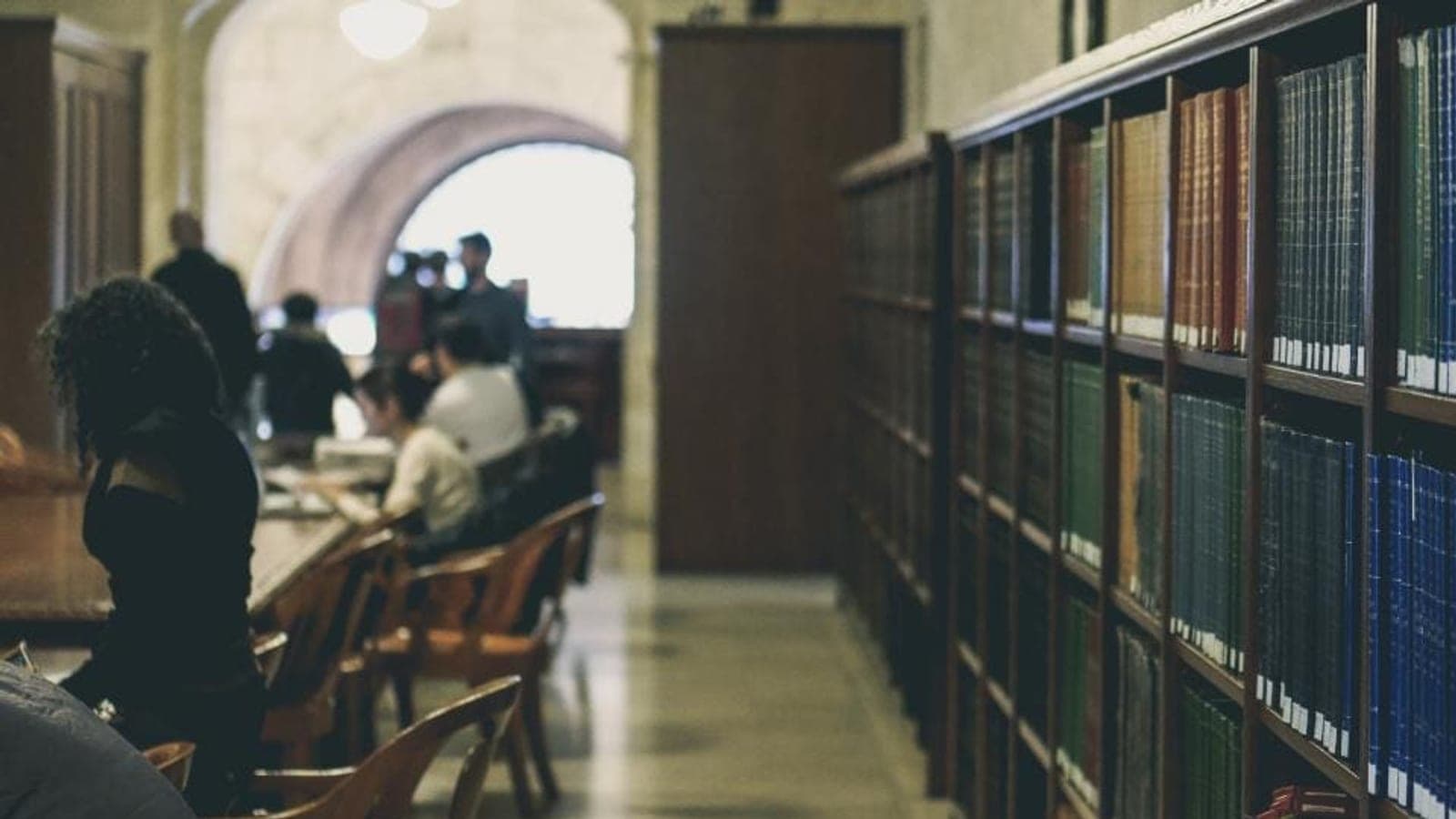 Une bibliothèque aux longues rangées de livres reliés, avec des étudiants assis à des tables en arrière-plan
