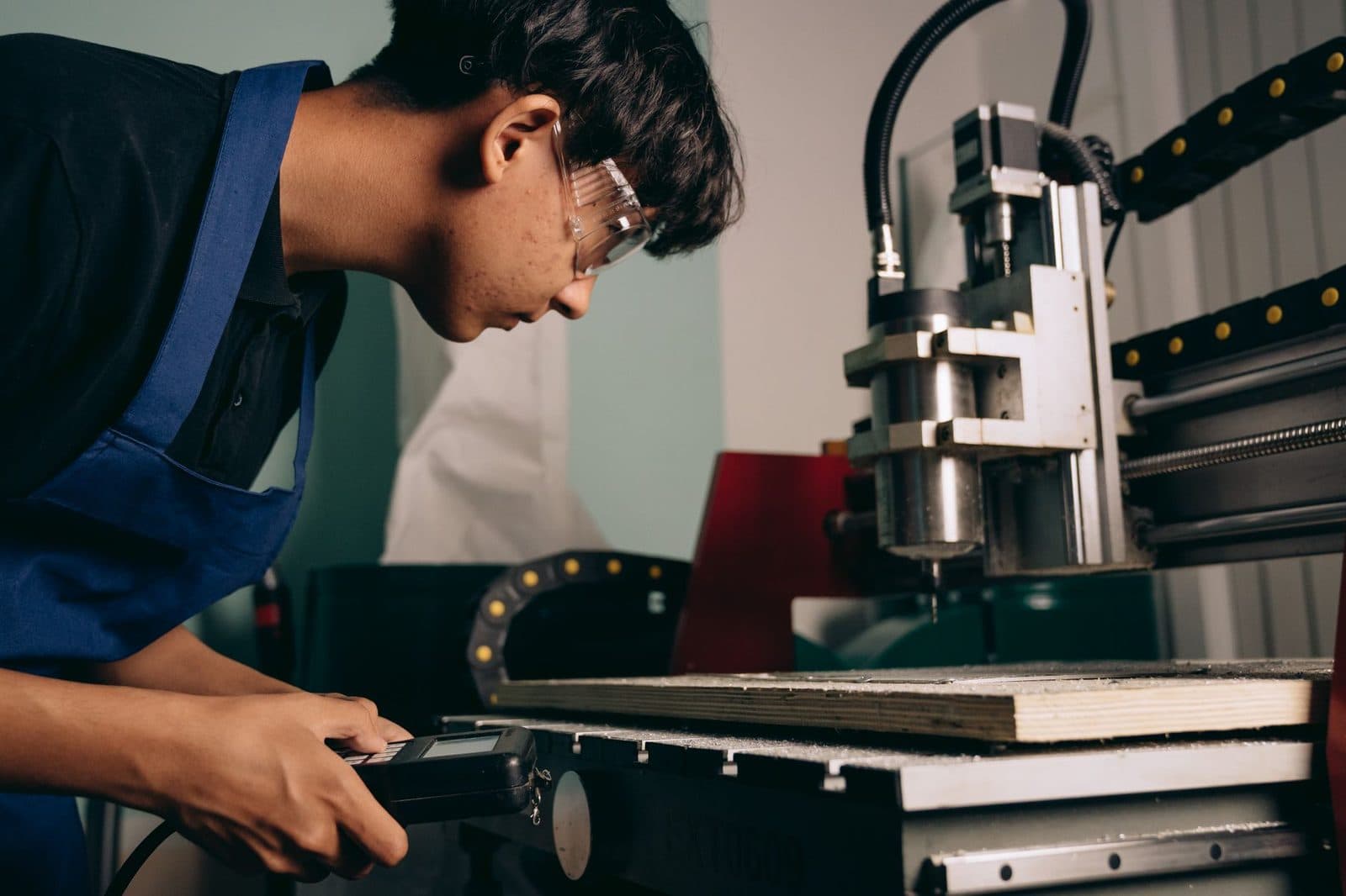 Young apprentice in blue overalls and safety goggles setting up a CNC machine in a workshop