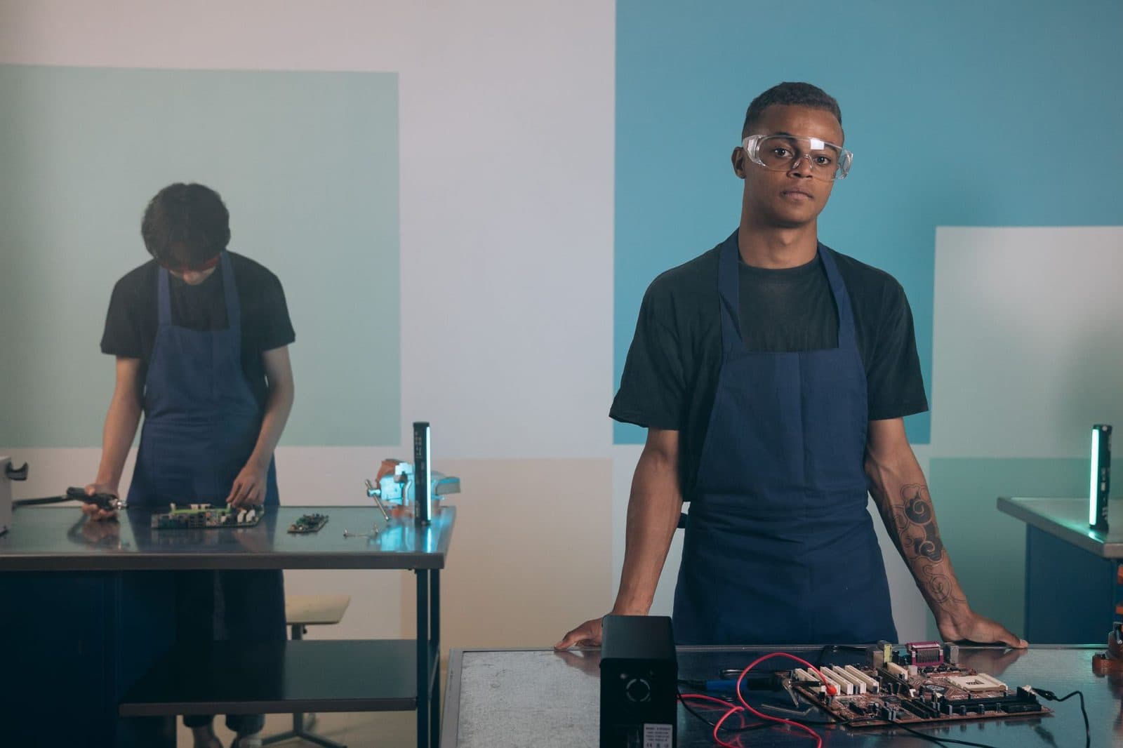 Deux jeunes apprentis en tablier bleu et lunettes de protection travaillant sur des cartes électroniques en atelier