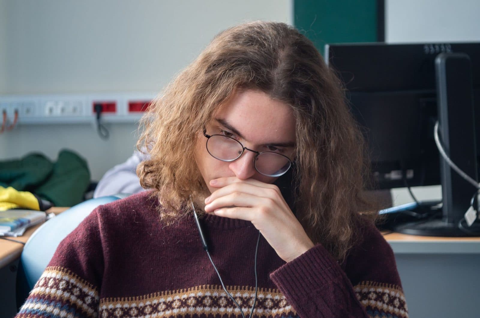 Jeune homme aux cheveux longs et bouclés, portant des lunettes et un pull en laine, pensif devant un écran d'ordinateur.