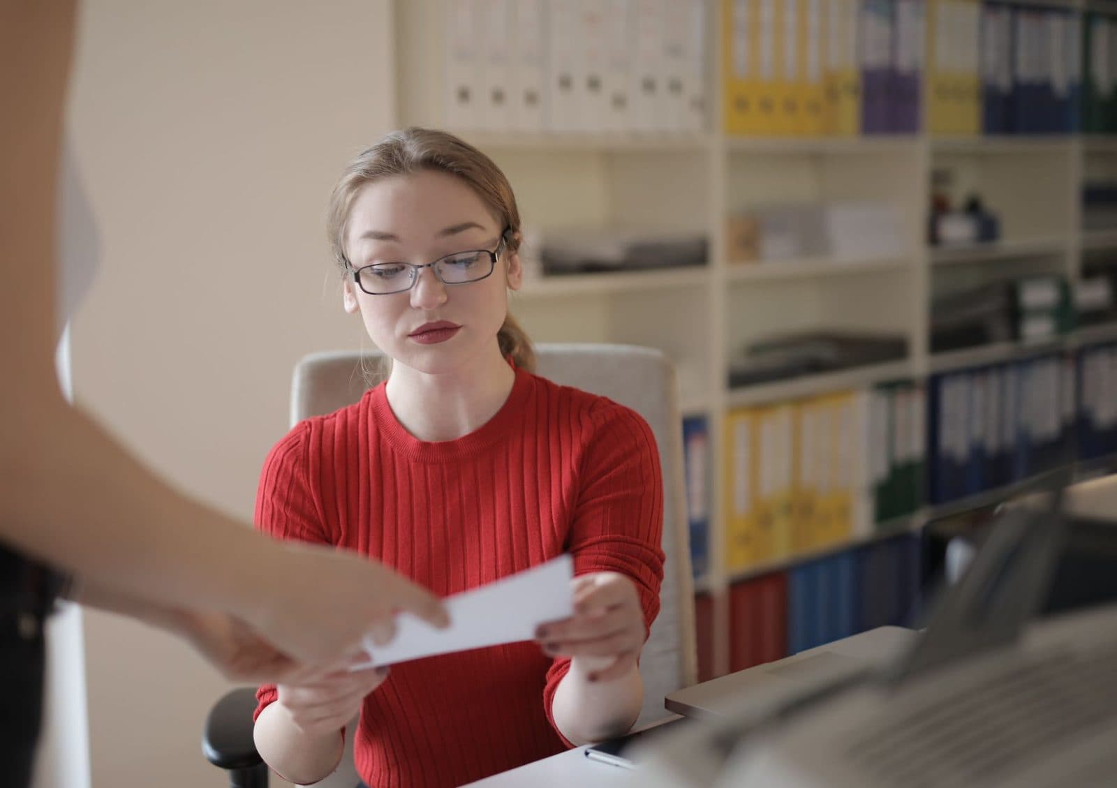 Jeune femme à lunettes en pull rouge recevant un document dans un bureau avec étagères de classeurs