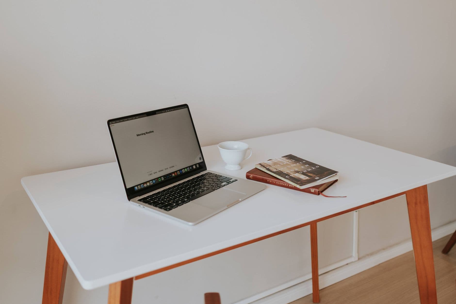 Un ordinateur portable ouvert, une tasse blanche et un carnet posés sur un bureau blanc.