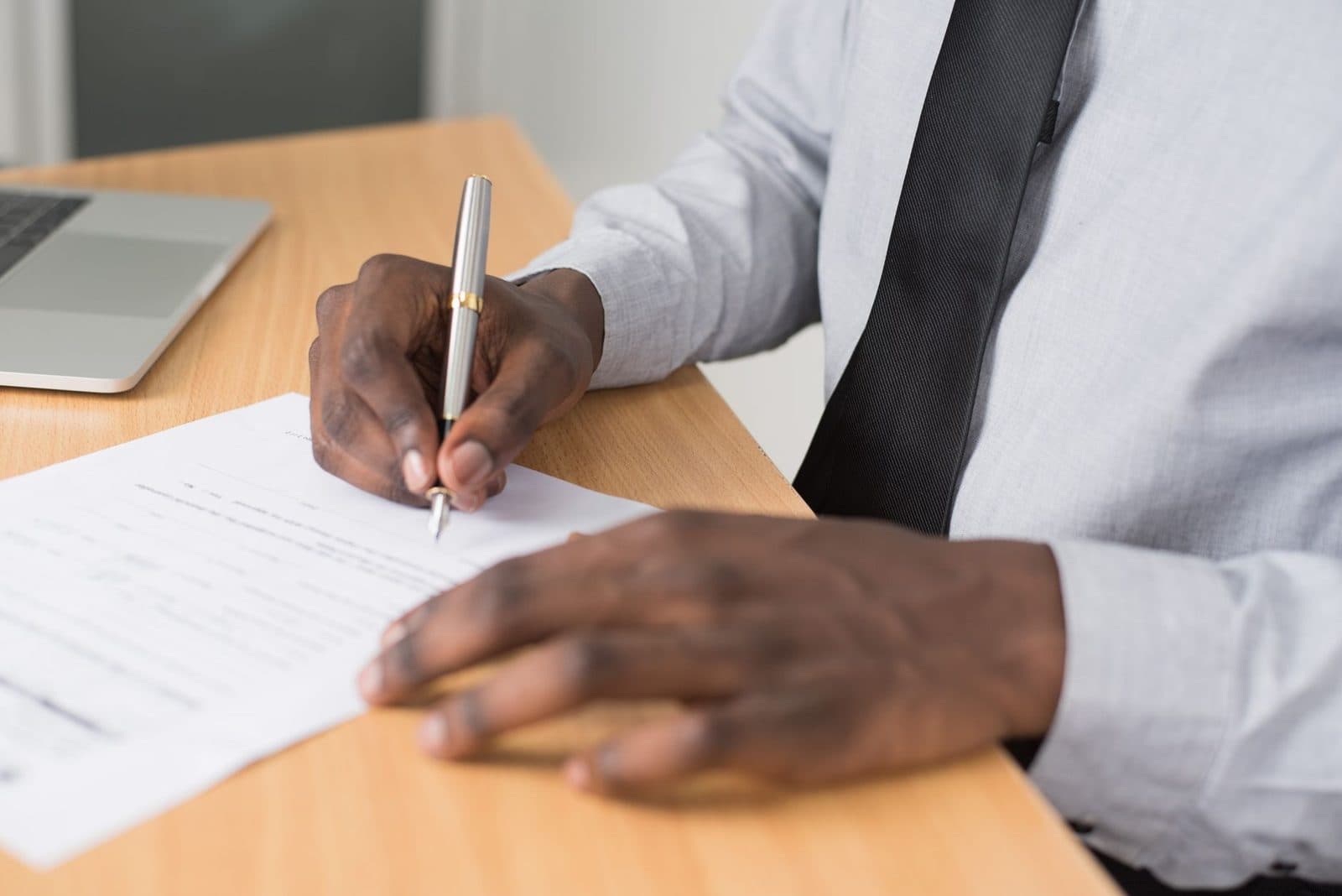Hombre con camisa y corbata firmando un documento en papel sobre un escritorio, junto a un ordenador portátil
