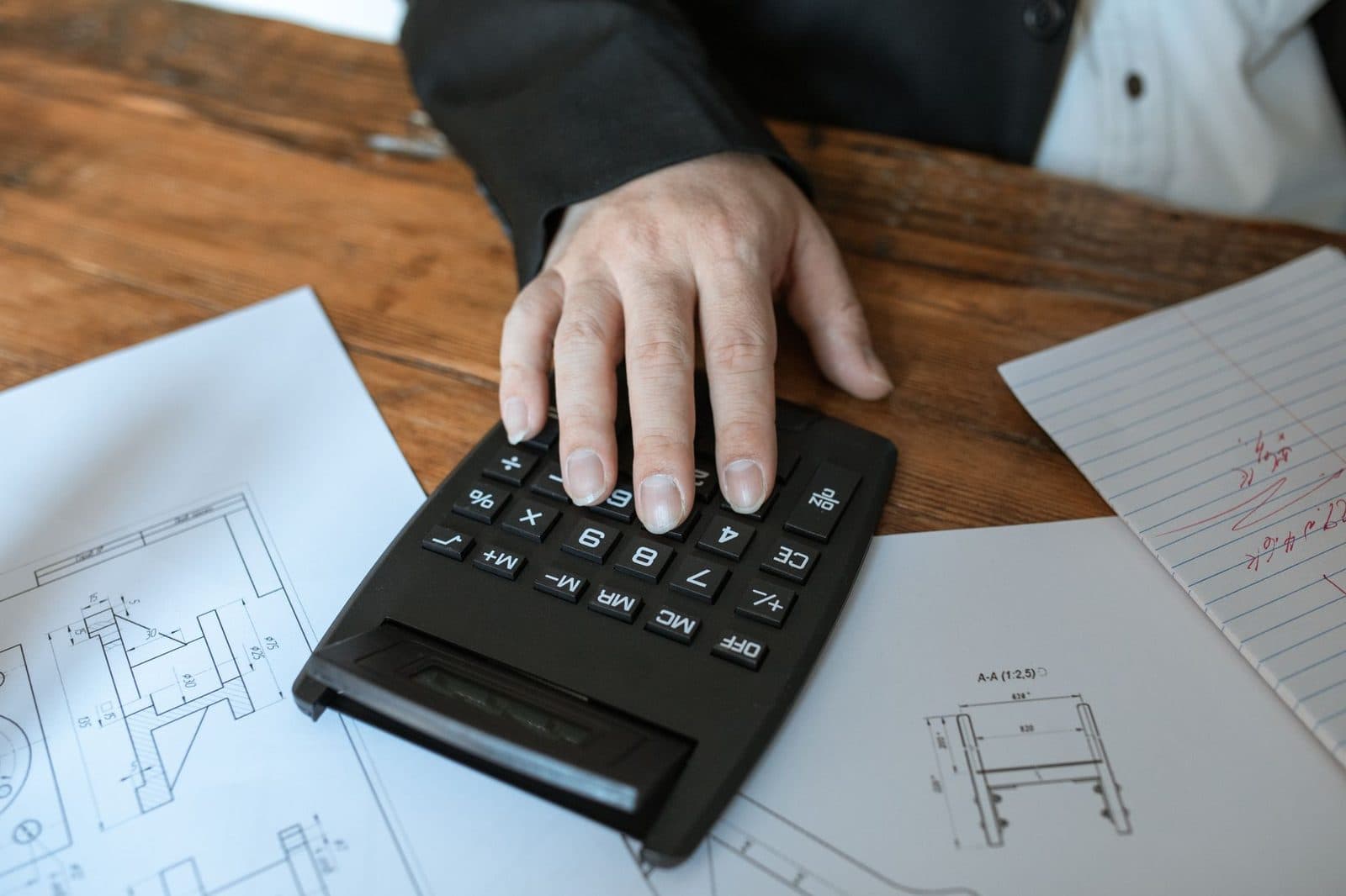 Main d'une personne en costume utilisant une calculatrice sur un bureau en bois avec des plans techniques