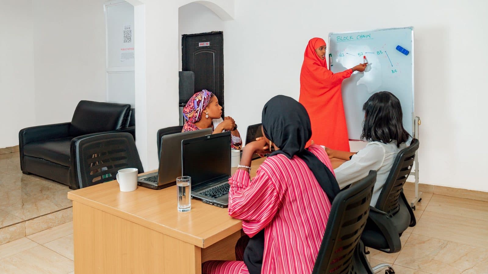 Une femme en hijab rouge présente un schéma sur un tableau blanc à trois autres femmes assises à un bureau.