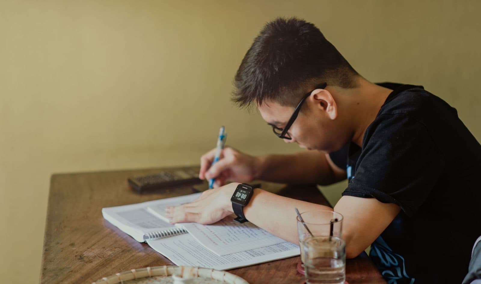 Un étudiant portant des lunettes écrit dans un cahier avec une calculatrice et un verre d'eau sur la table.