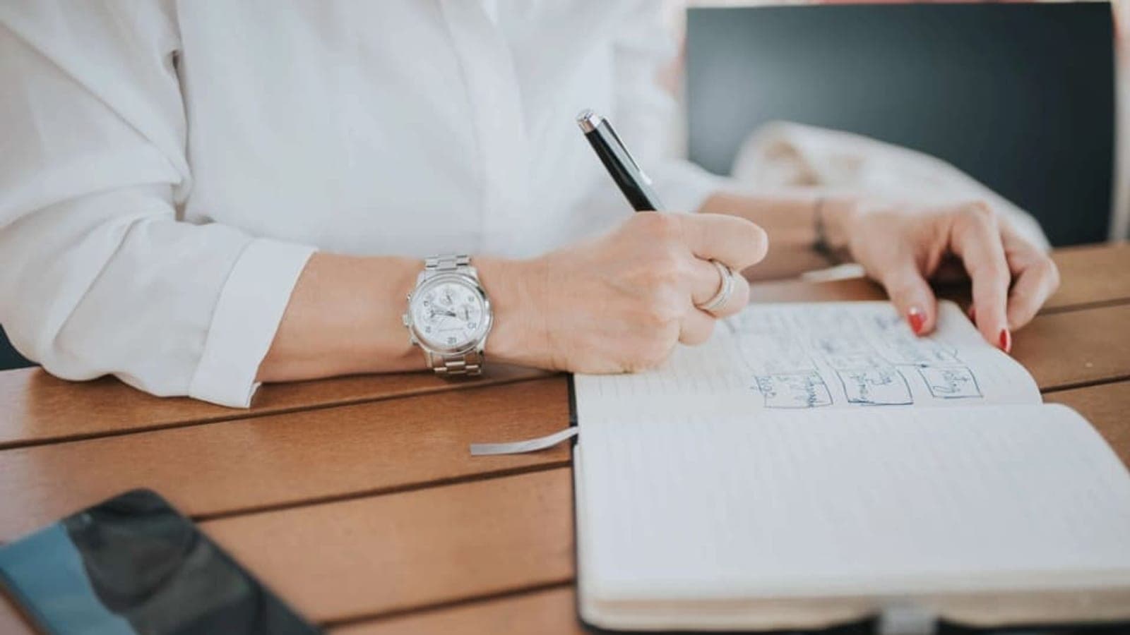 Une personne en chemise blanche, montre au poignet, écrit au stylo dans un carnet ouvert posé sur une table en bois, à côté d'un téléphone