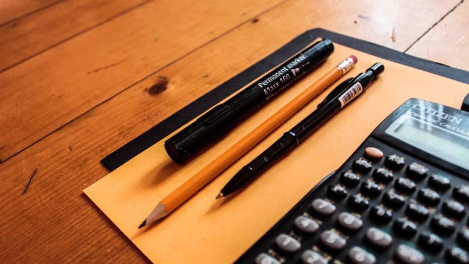 Une calculatrice, un crayon, un stylo et un marqueur posés sur une chemise cartonnée orange, sur une table en bois
