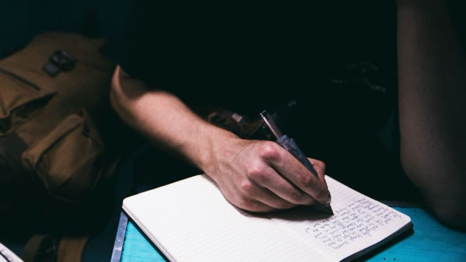 Une main écrit au stylo dans un carnet ouvert posé sur une table bleue, à côté d'un sac à dos