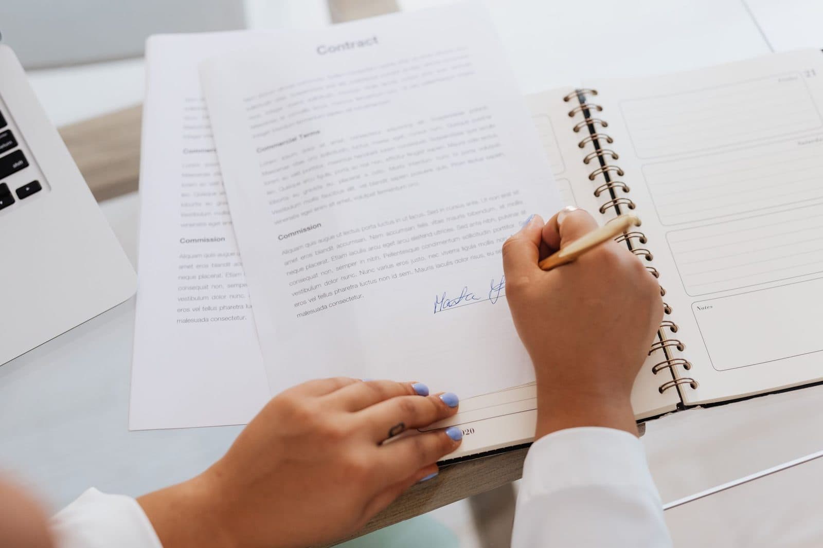 Jeune femme signant un contrat posé sur un bureau, à côté d'un agenda ouvert et d'un ordinateur portable