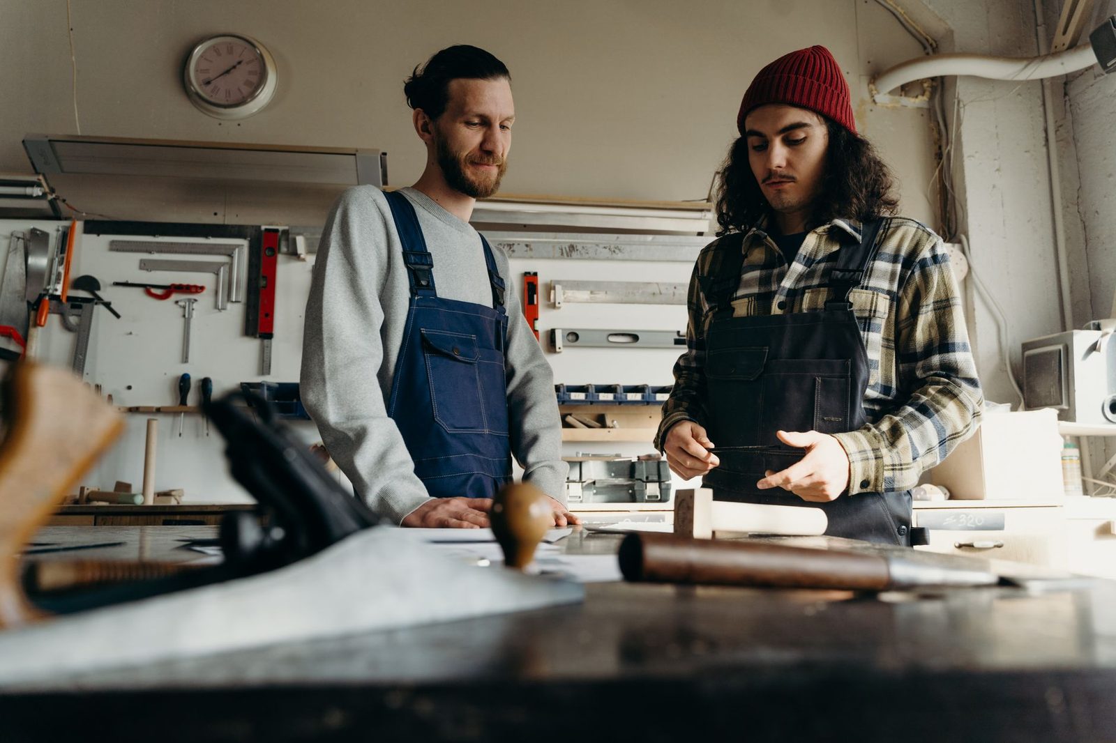 Deux artisans en salopette discutent devant un établi dans un atelier de menuiserie équipé d'outils