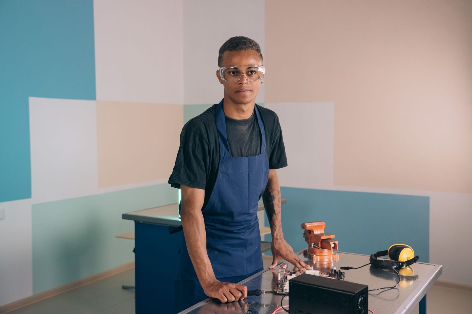 Jeune homme en tablier bleu et lunettes de protection dans un atelier, devant une carte électronique et un étau