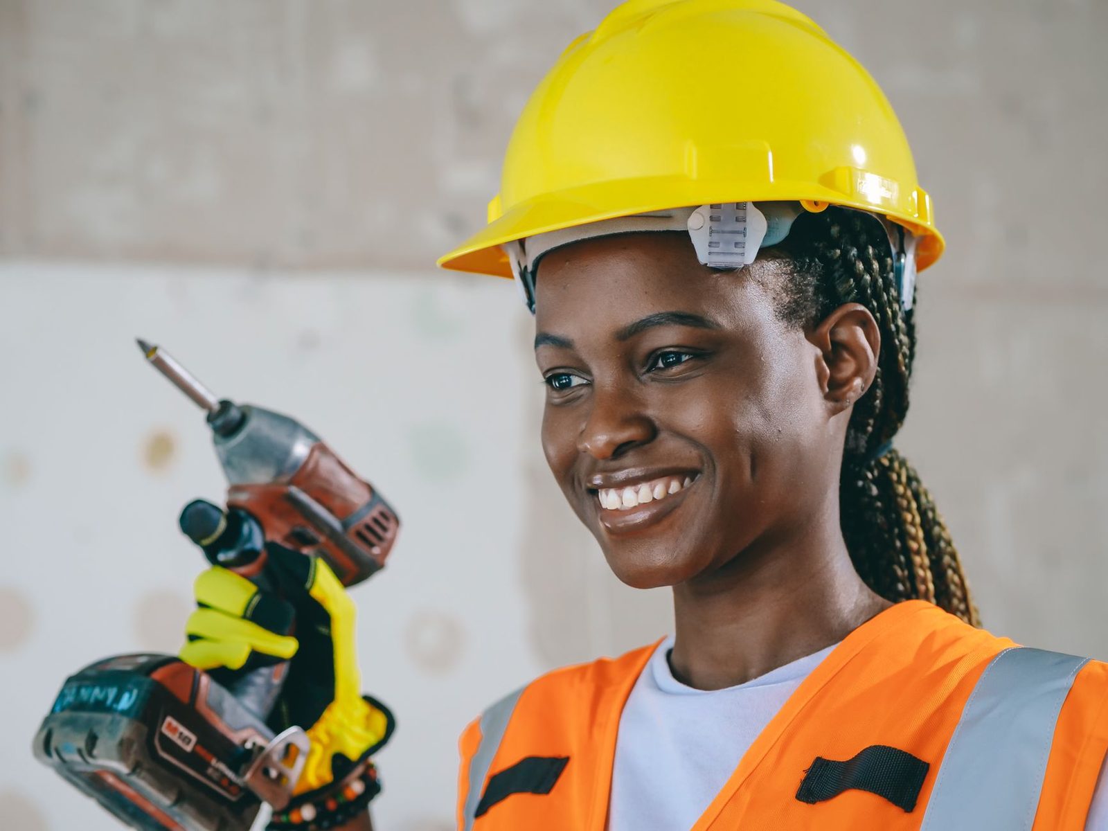 Jeune femme souriante en casque jaune et gilet orange tenant une visseuse sur un chantier
