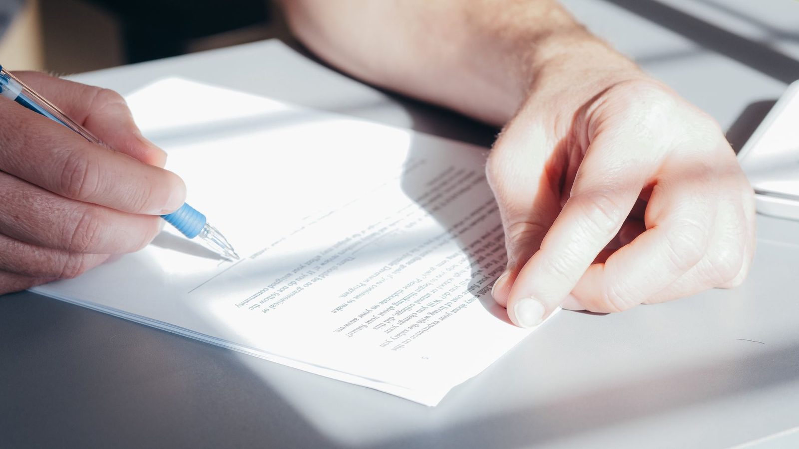 Hands signing a paper document with a blue pen on a light-coloured desk