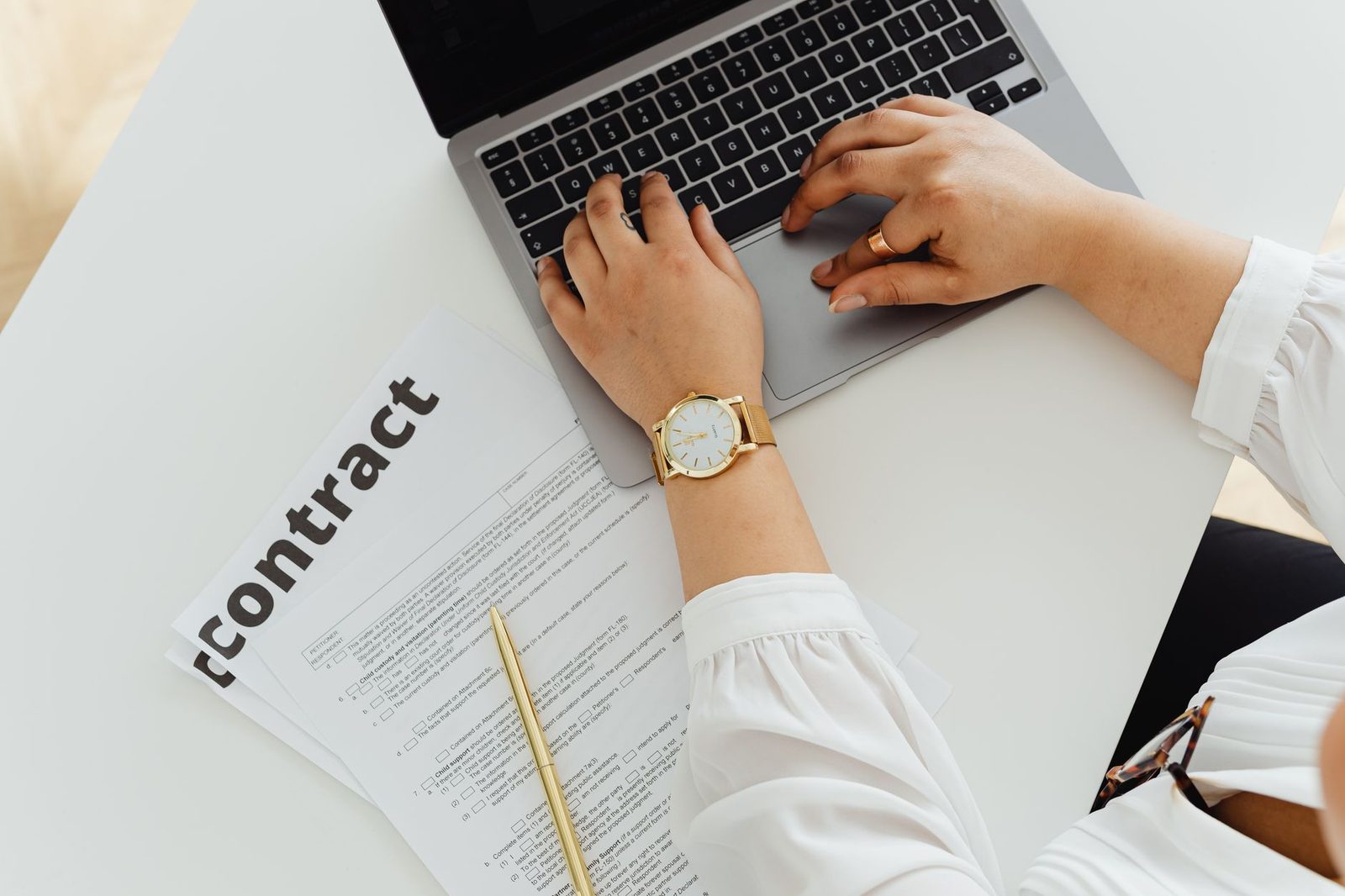 Woman in a white blouse typing on a laptop, next to a paper contract and a gold pen