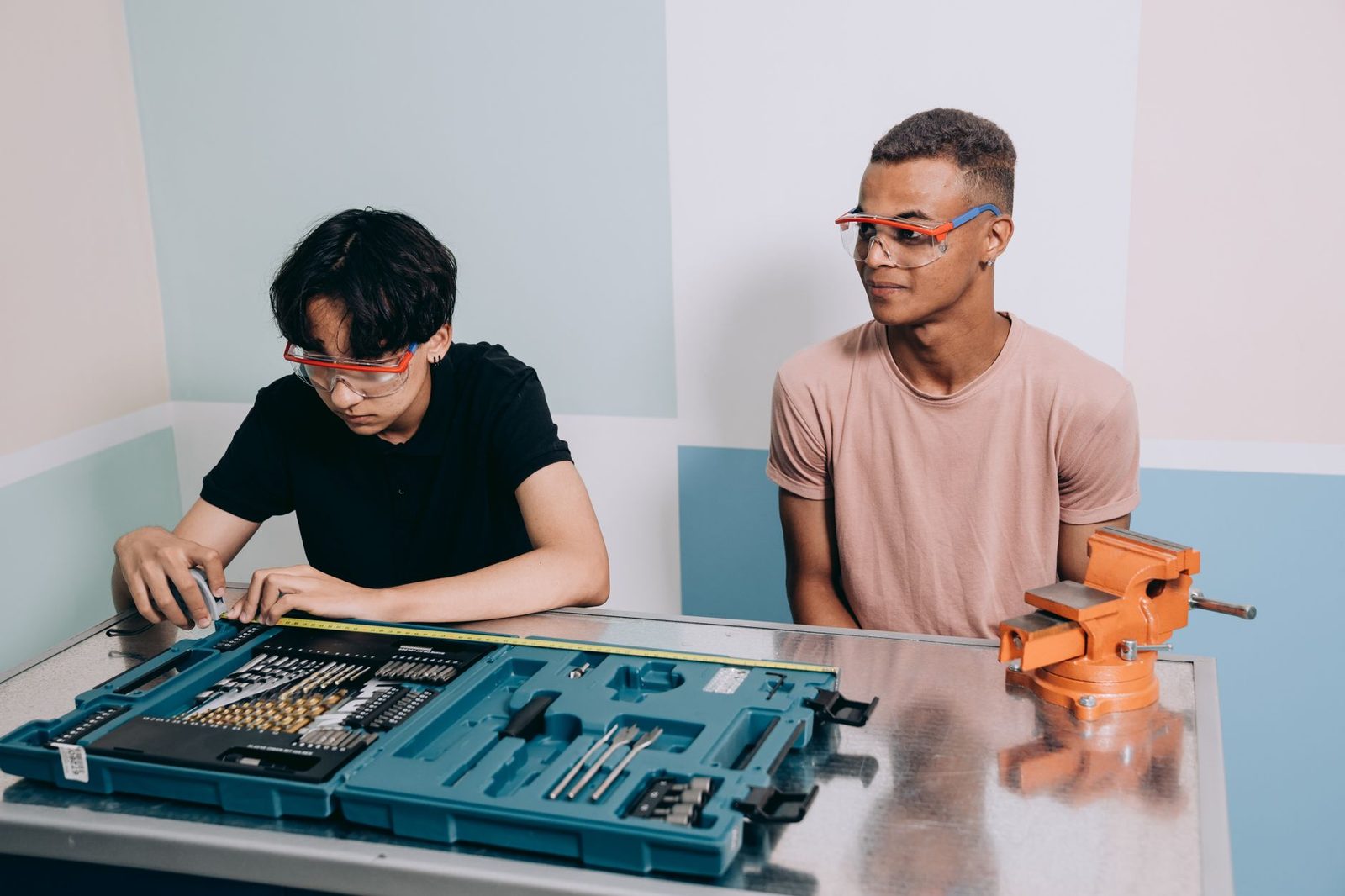 Deux jeunes en atelier avec lunettes de protection, coffret d'outils, mètre ruban et étau orange