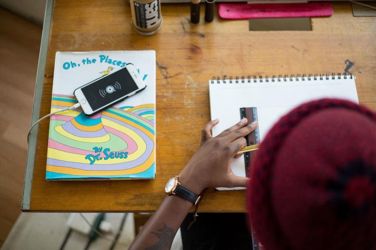 Vue de dessus d'un bureau en bois : une personne trace un trait à la règle sur un carnet à spirale, à côté d'un livre et d'un téléphone
