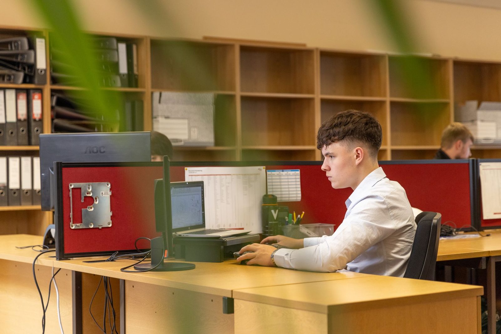 Jeune homme en chemise blanche travaillant devant un ordinateur dans un bureau avec étagères à classeurs
