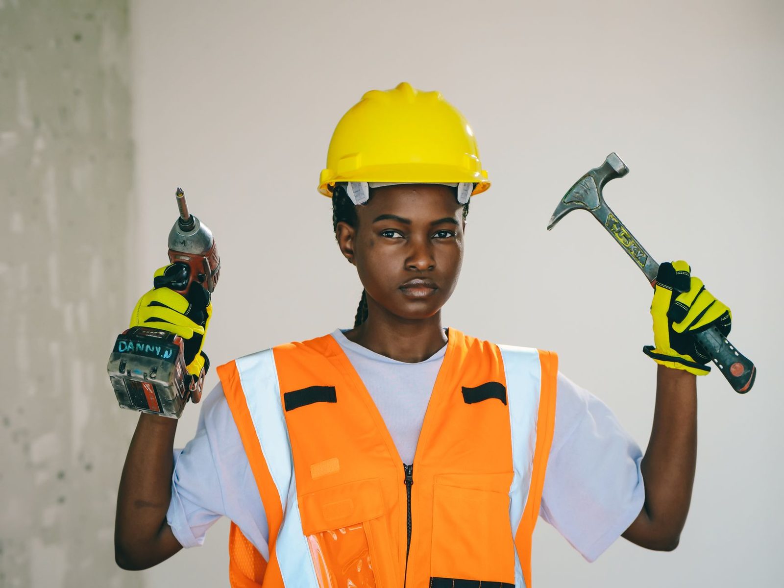 Jeune femme en gilet orange et casque jaune tenant une perceuse et un marteau sur un chantier