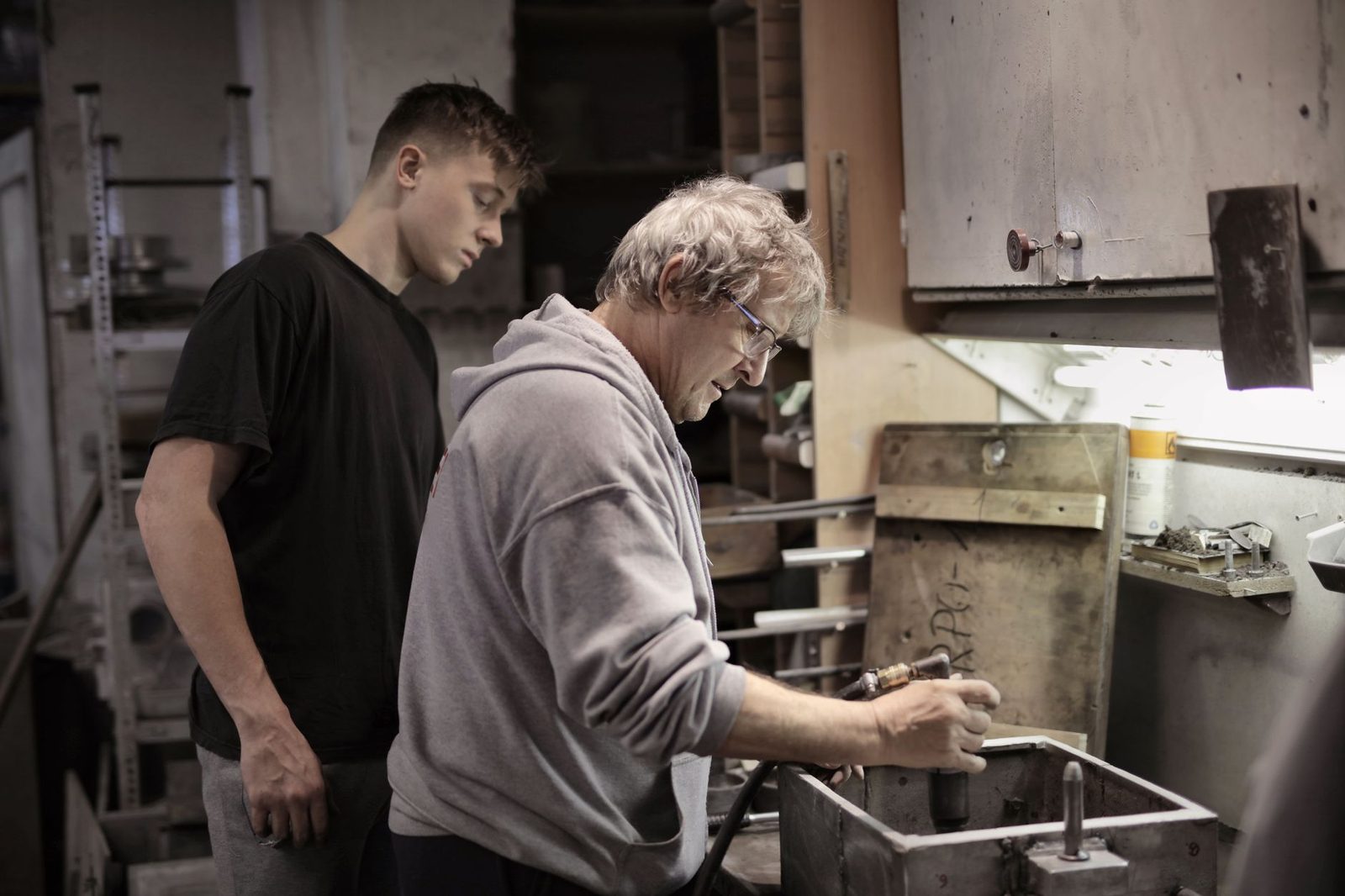 Un jeune apprenti observe un artisan expérimenté travaillant sur une pièce métallique dans un atelier