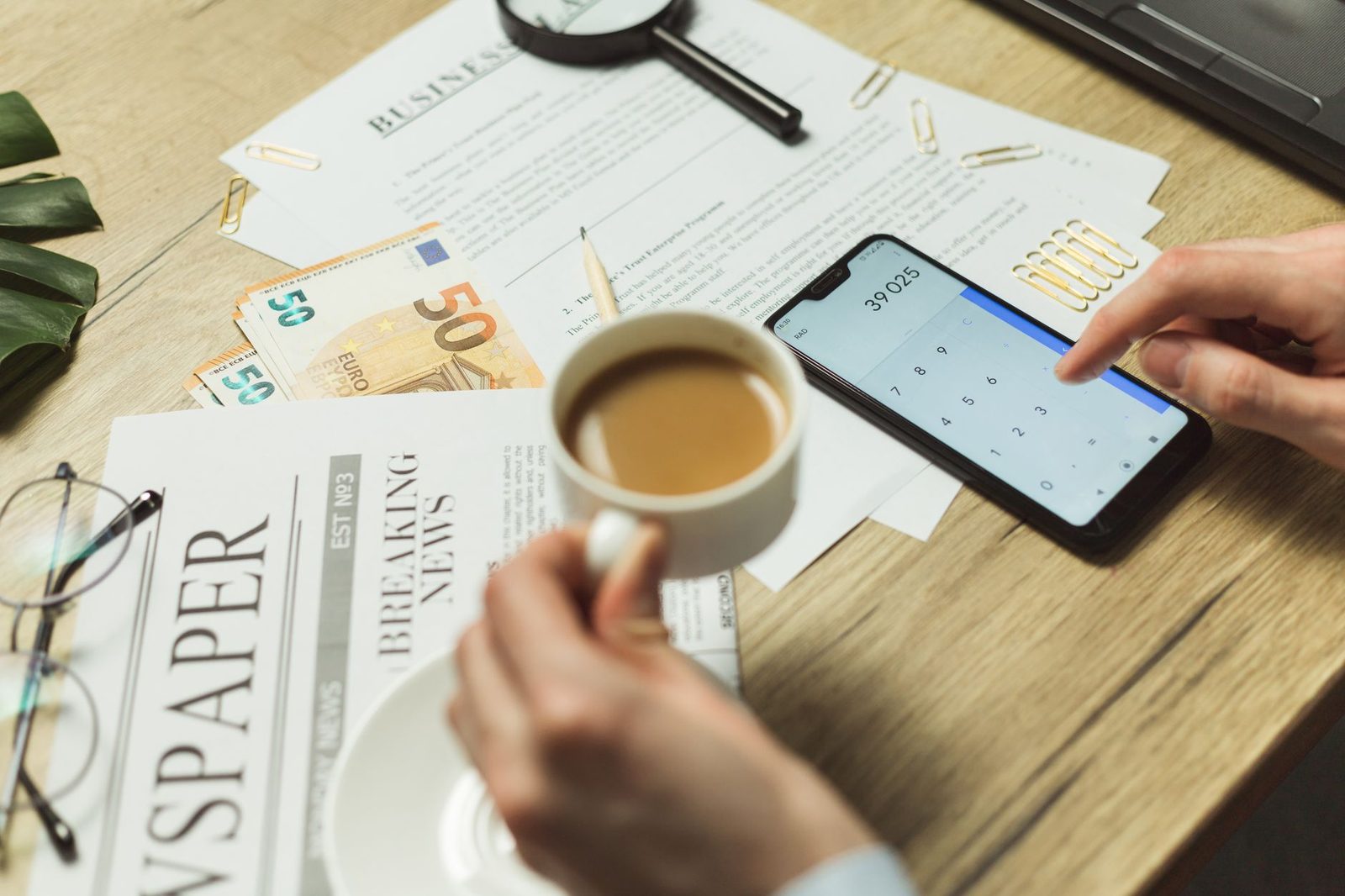 Personne calculant sur un smartphone devant des billets en euros, des documents et une tasse de café