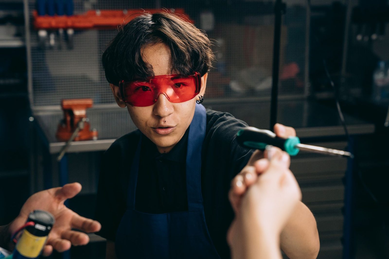 Jeune apprenti en tablier bleu et lunettes de protection rouges recevant un tournevis dans un atelier