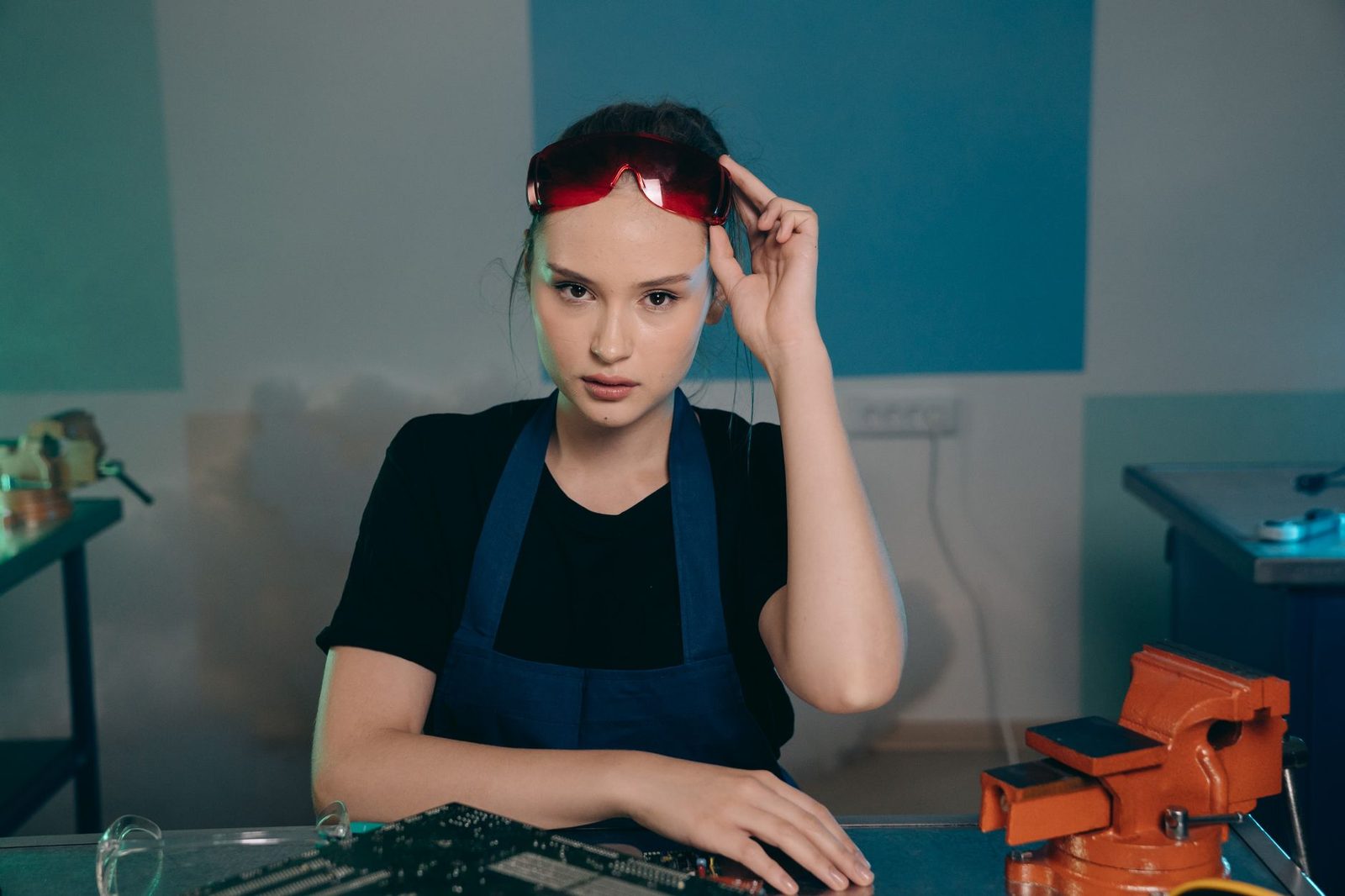Jeune femme en tablier bleu avec lunettes de protection dans un atelier, près d'un étau et d'une carte électronique