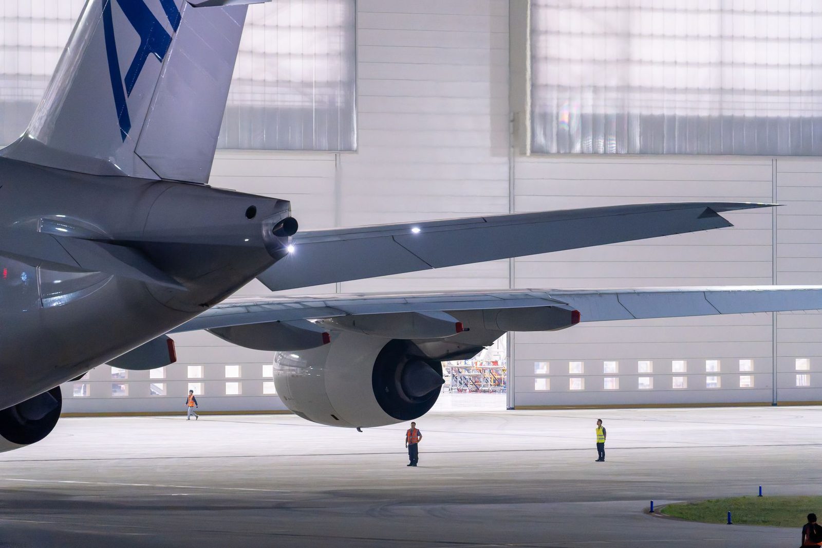 Empennage et aile d'un avion devant un hangar, avec des techniciens en gilets fluorescents sur le tarmac