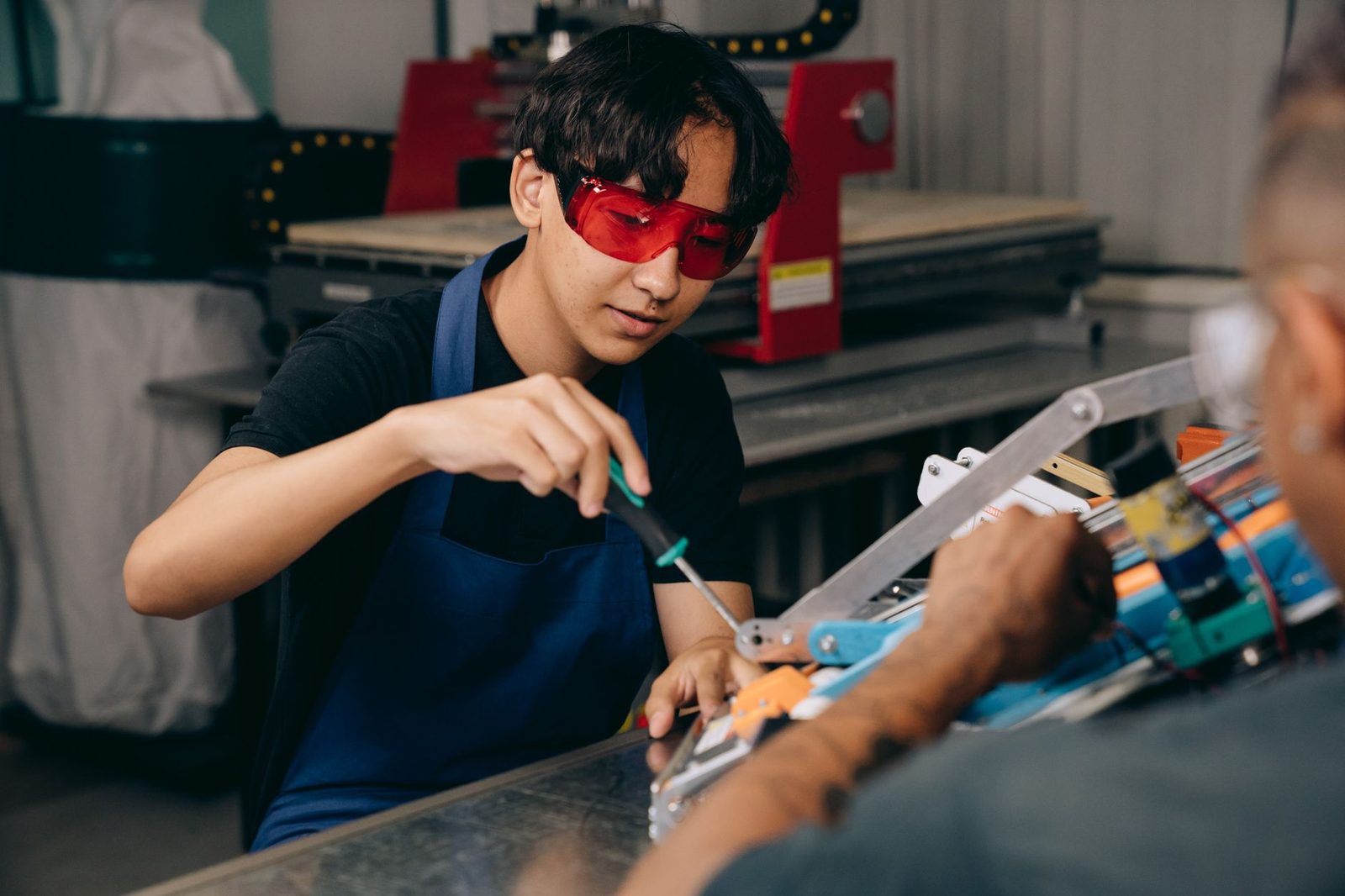 Jeune apprenti avec lunettes de protection et tablier bleu assemblant une pièce en atelier avec un formateur