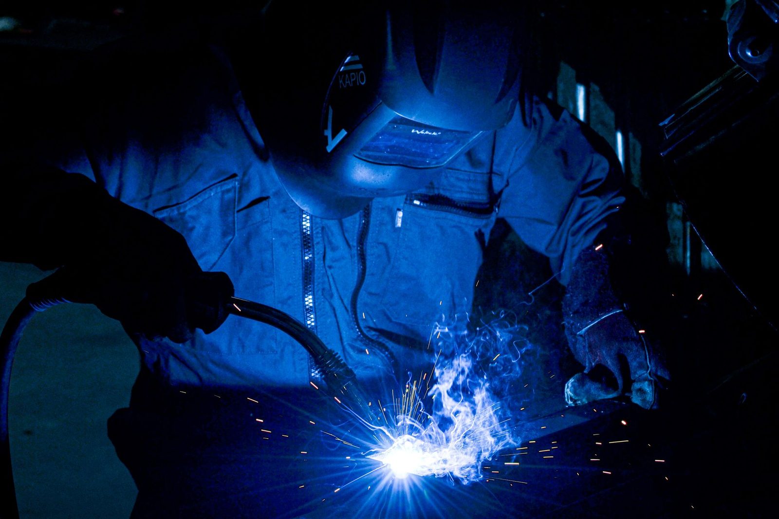 Soudeur casqué en bleu de travail réalisant une soudure à l'arc, gerbe d'étincelles dans l'atelier