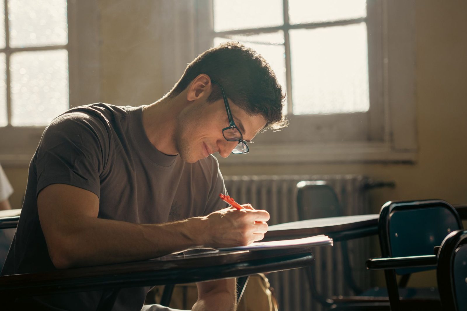 Un étudiant portant des lunettes écrit sur un cahier à son bureau dans une salle de classe.