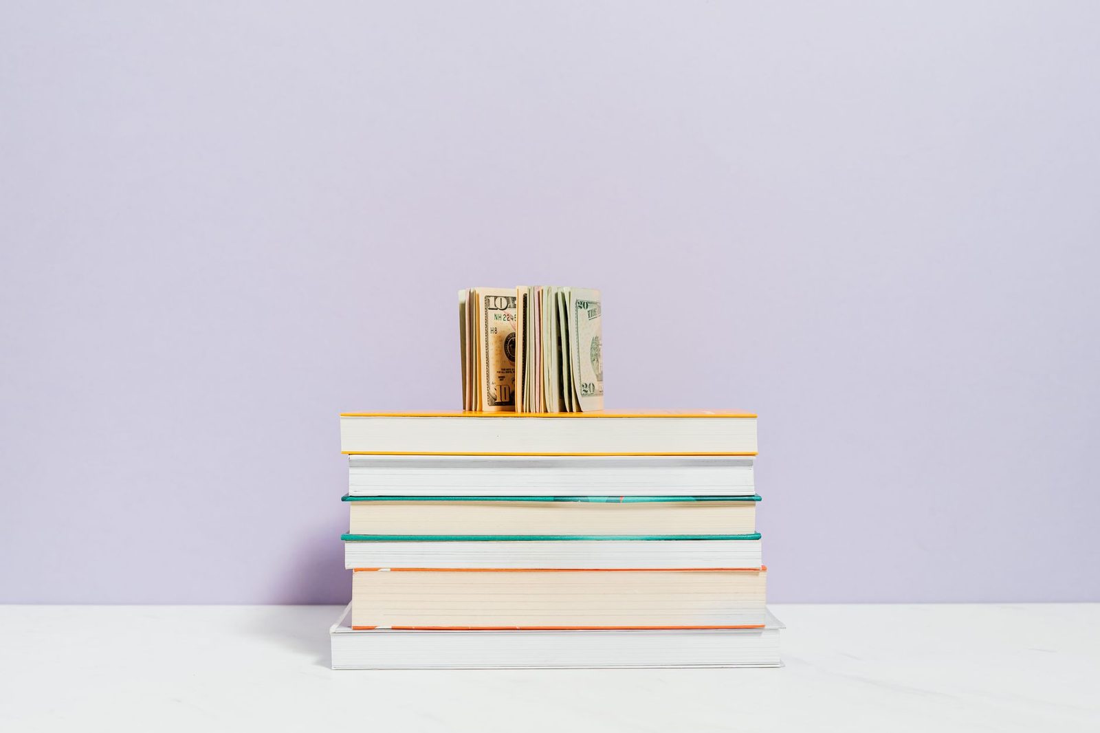 Une pile de livres avec des billets de banque américains posés sur le dessus, sur un fond violet.
