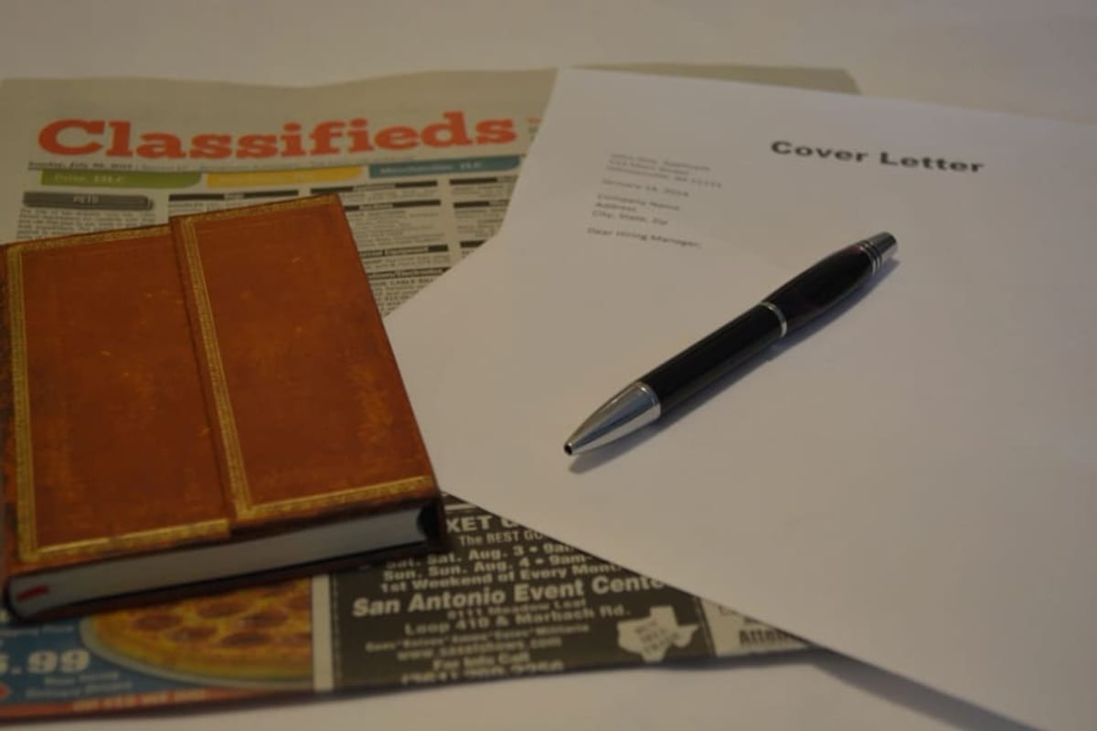 Une lettre de motivation et un stylo posés sur un journal de petites annonces, à côté d'un carnet relié en cuir