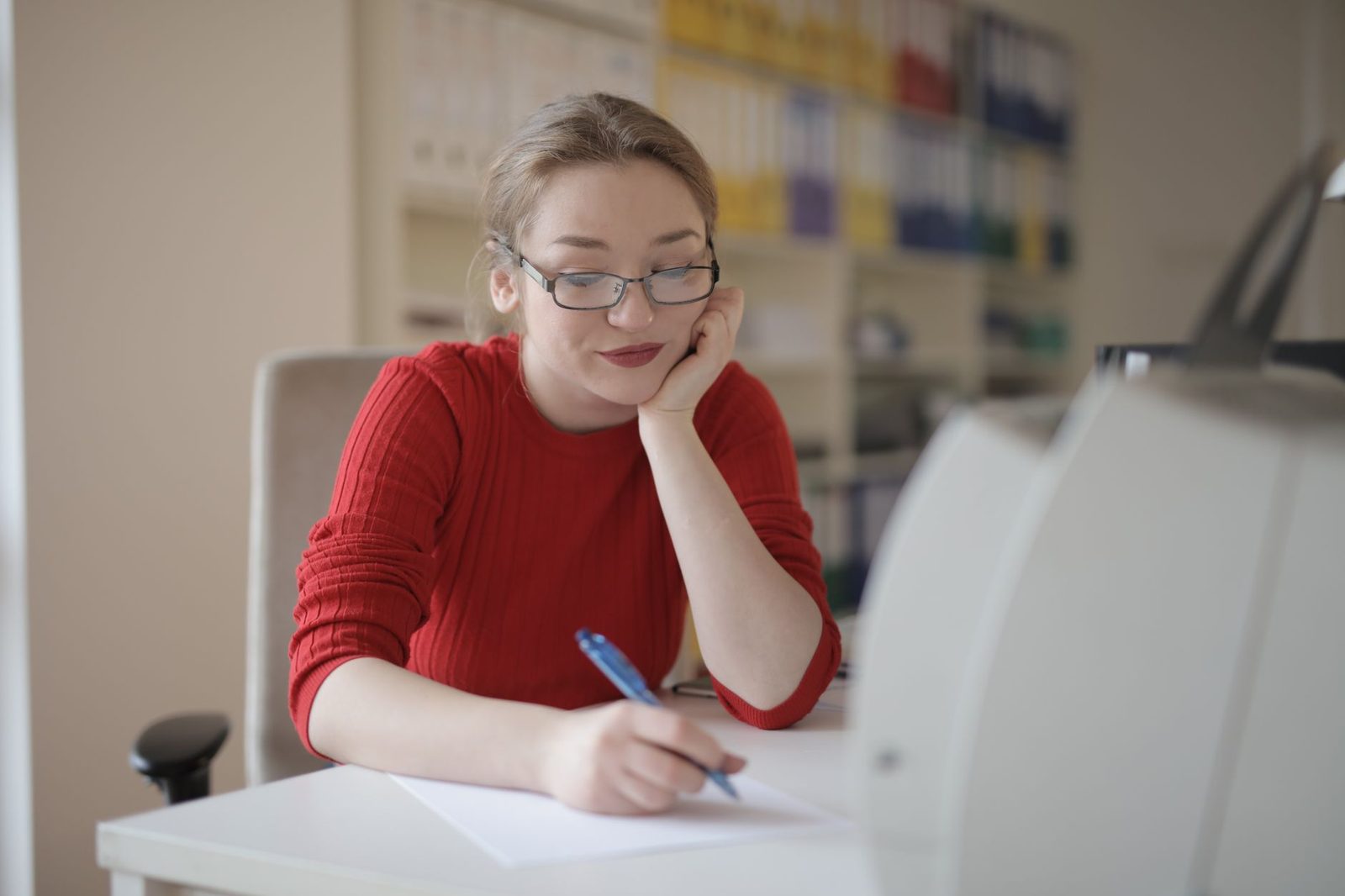Une jeune femme avec des lunettes, portant un pull rouge, écrit sur une feuille de papier devant un ordinateur.