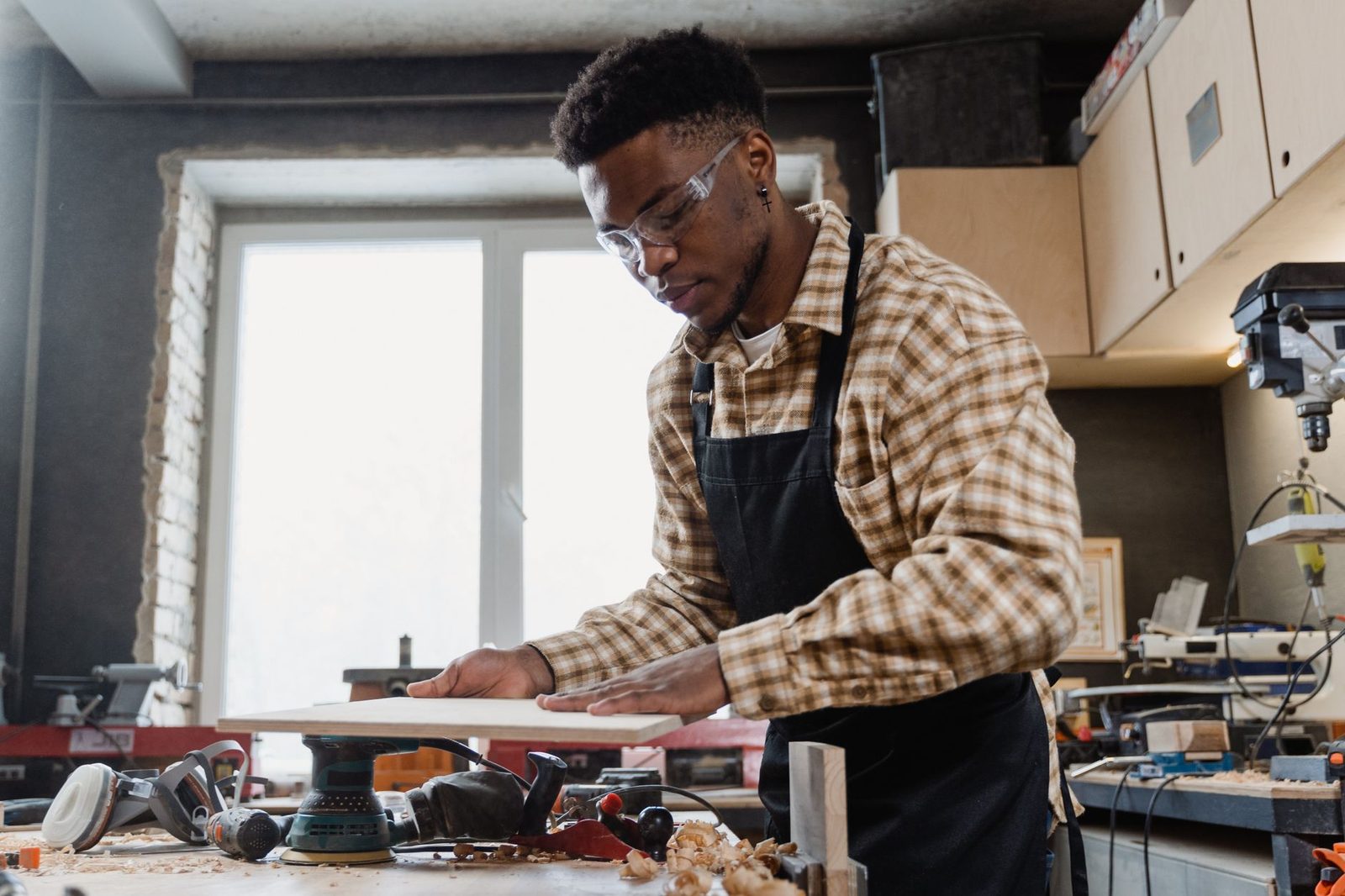 Jeune homme en tablier et lunettes de protection ponçant une planche de bois dans un atelier de menuiserie