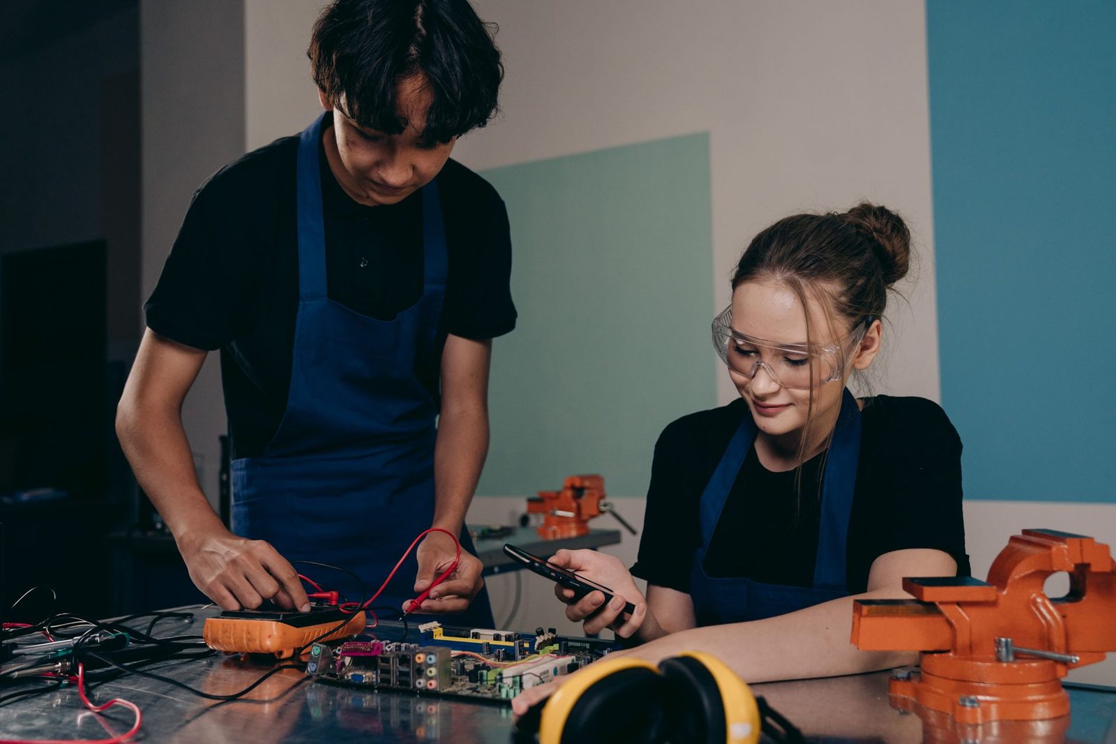Deux apprentis en tablier bleu testent une carte électronique avec un multimètre dans un atelier
