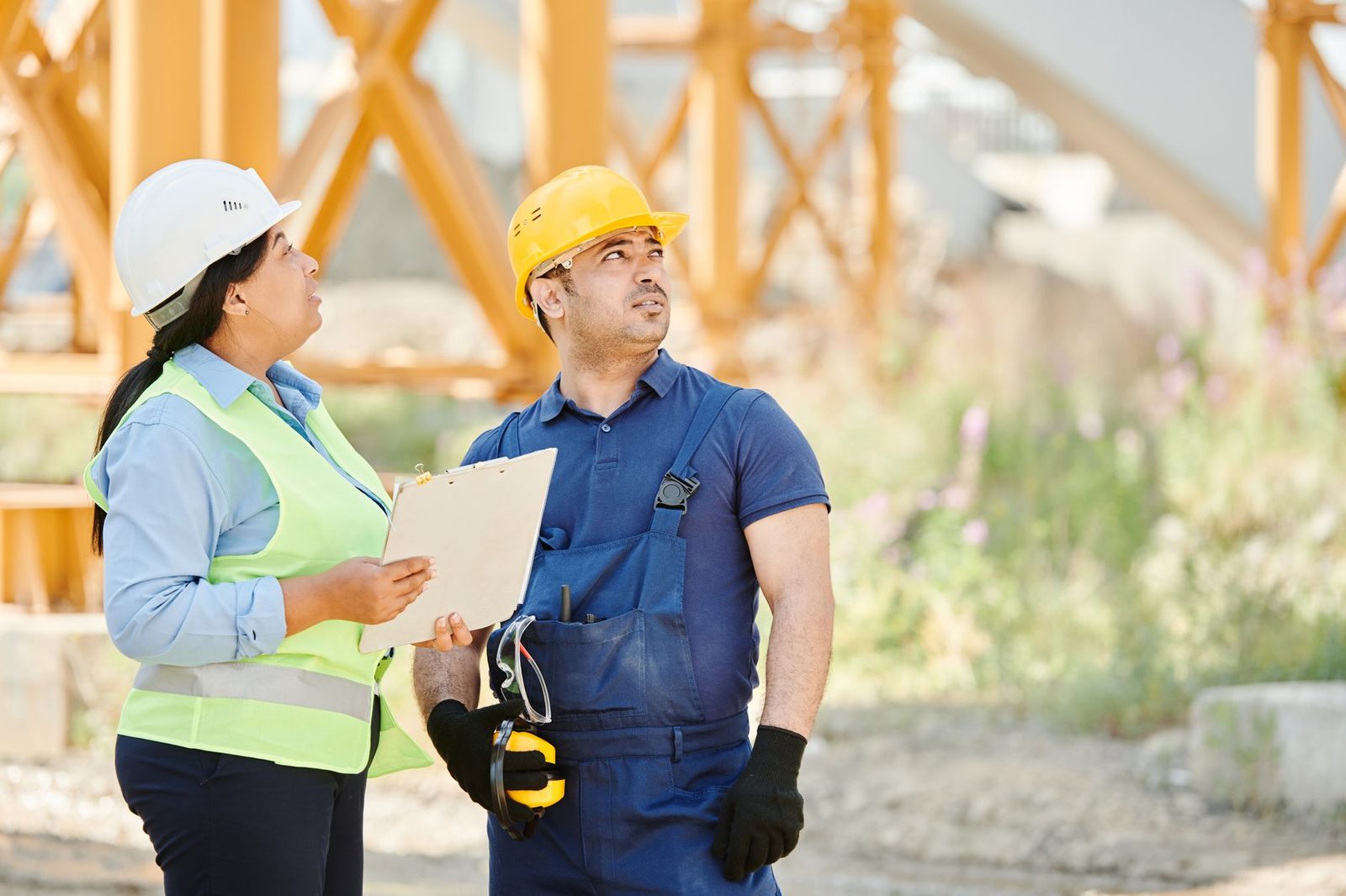 Femme avec casque blanc et gilet jaune tenant un porte-bloc face à un ouvrier casqué sur un chantier
