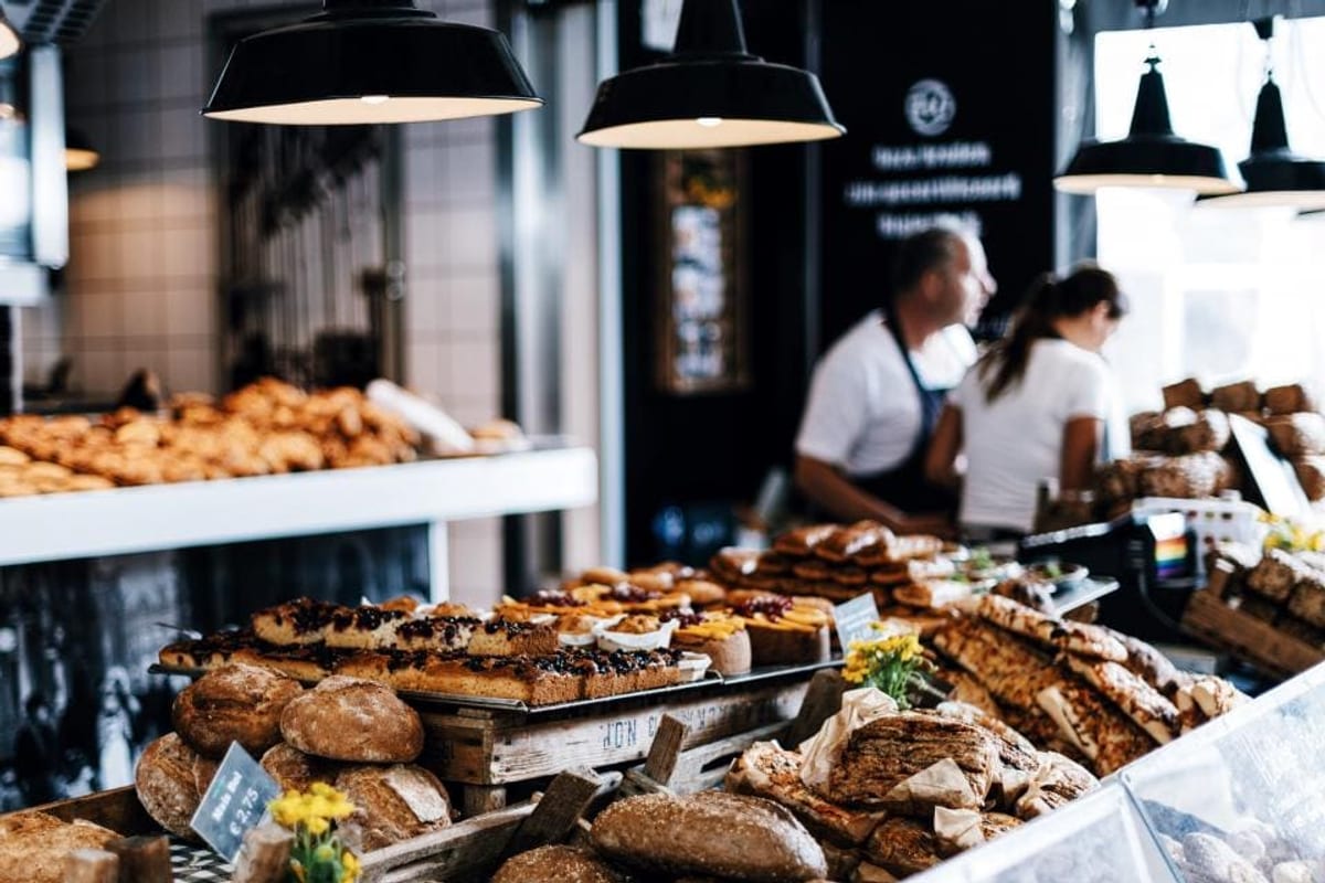 Comptoir d'une boulangerie chargé de pains et de pâtisseries, avec deux employés en tablier au second plan