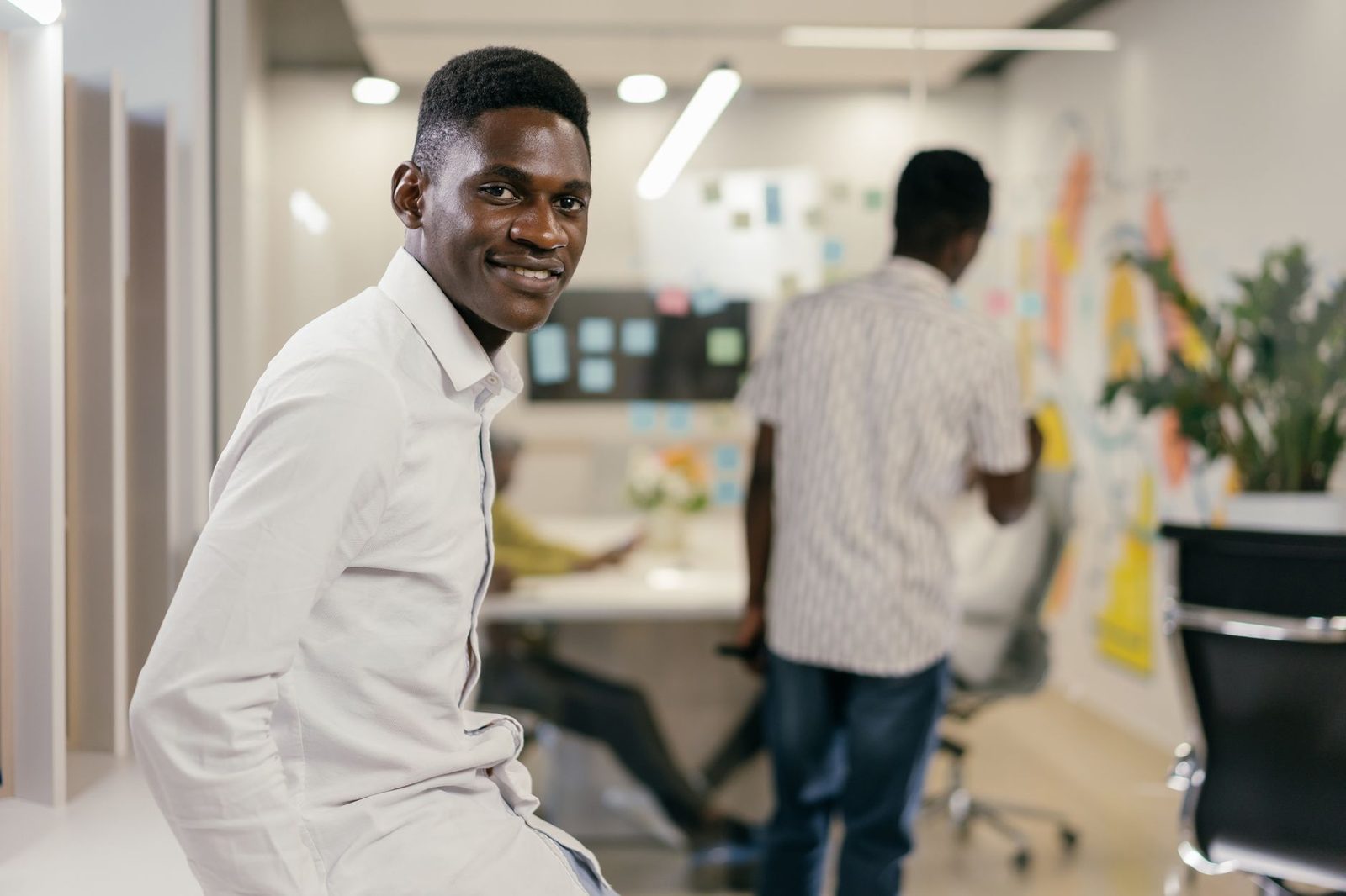 Jeune homme en chemise blanche souriant dans un bureau open space, collègues travaillant en arrière-plan