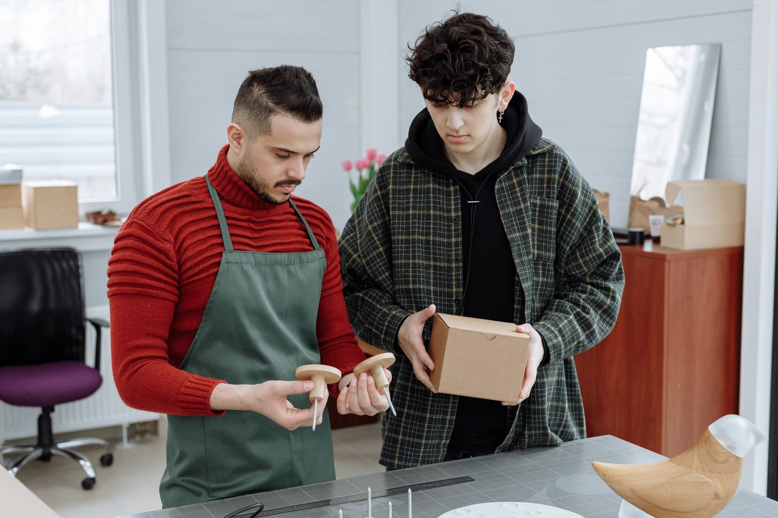 Un artisan en tablier montre une pièce en bois à un jeune apprenti tenant un carton dans un atelier