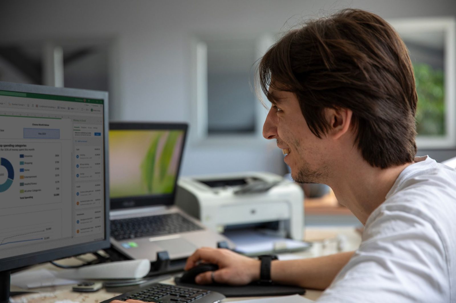 Jeune homme souriant travaillant au bureau devant un écran, un ordinateur portable et une imprimante